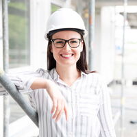 Eine Frau lehnt an einer Wand, sie trägt einen weißen Schutzhelm und lächelt.
